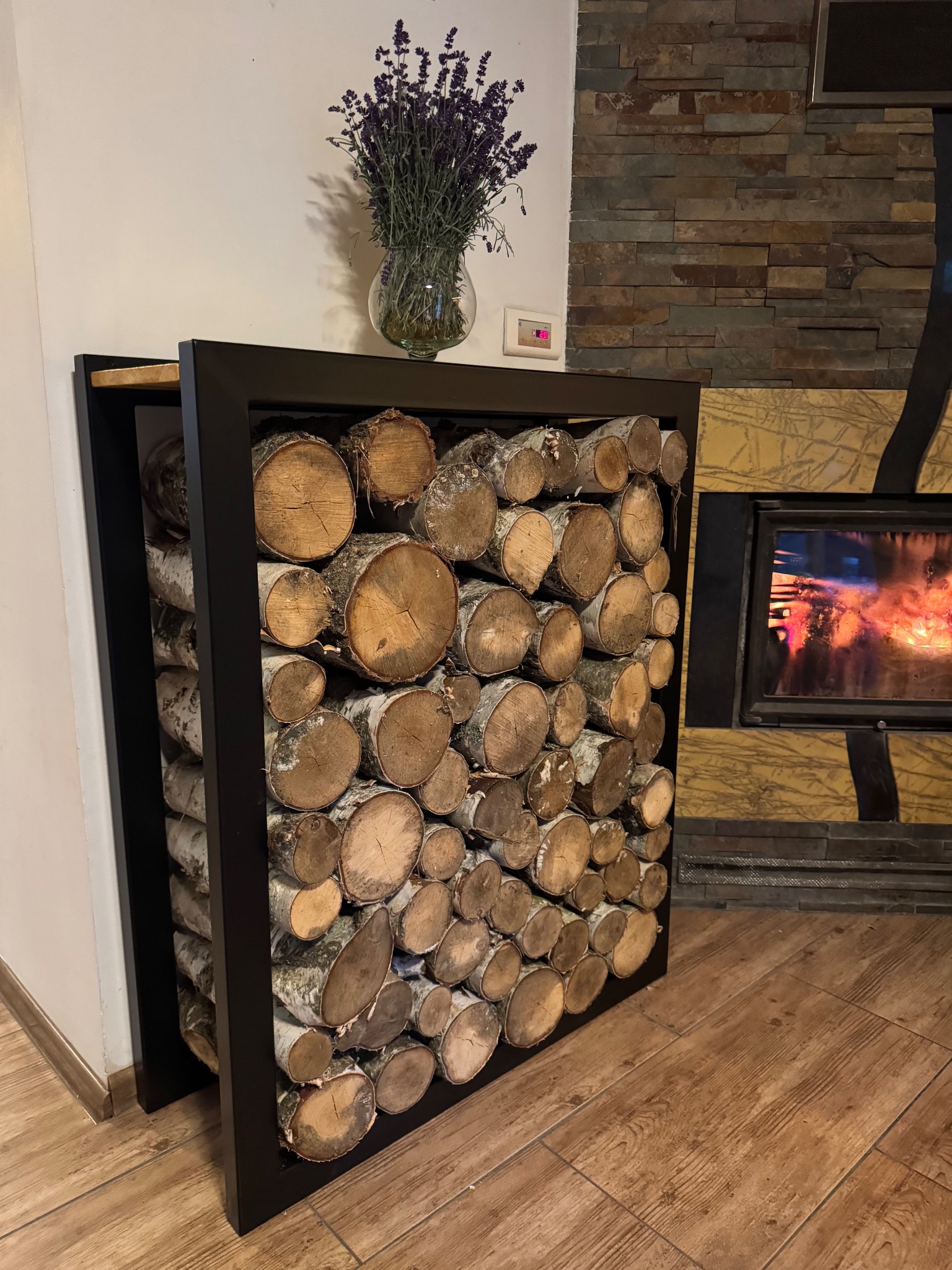Metalowy regał na drewno kominkowe z czarną ramą, wypełniony pociętymi brzozowymi klockami. Nad nim wazon z lawendą, w tle kominek z paleniskiem.