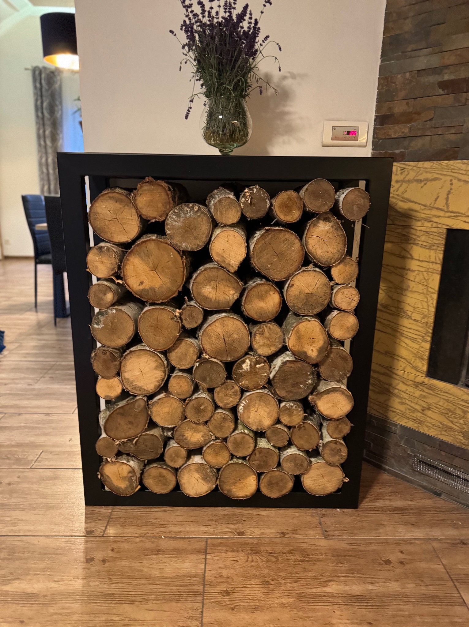 Metalowa rama wypełniona drewnem kominkowym, tworząca dekoracyjny element wnętrza. U góry wazon z lawendą. Obok widoczny fragment obudowy kominka.