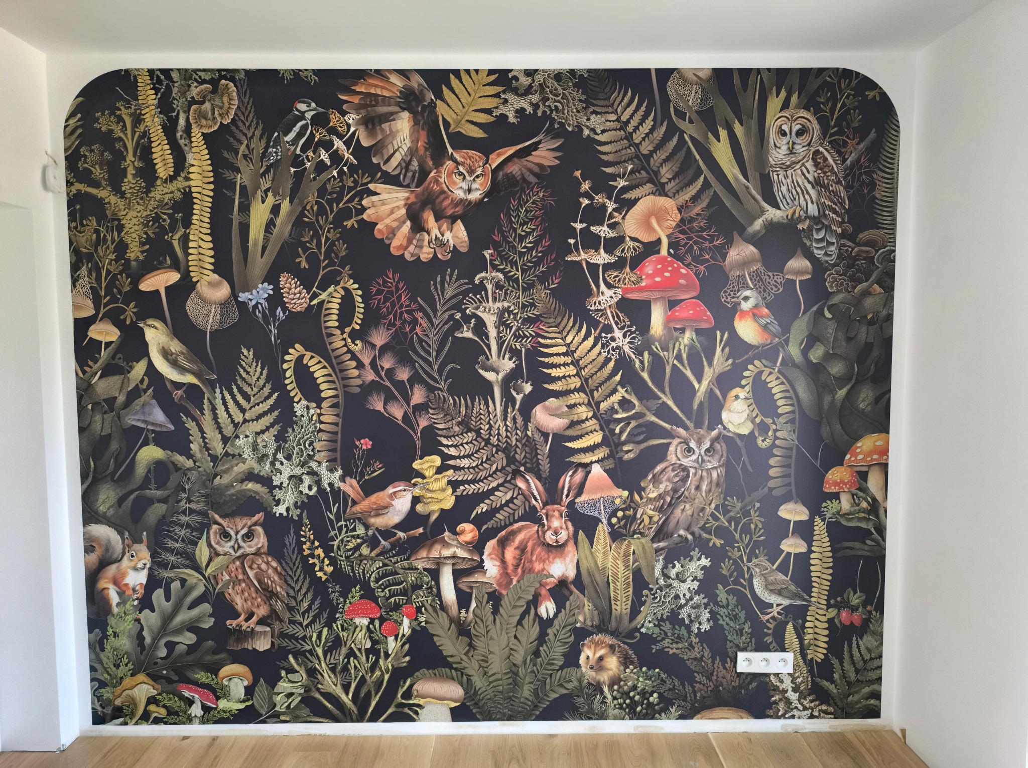 Ściana po dekoracji tapetą z motywem leśnym: sowy, grzyby, króliki i rośliny na ciemnym tle. Widoczne gniazdka elektryczne i drewniana podłoga.