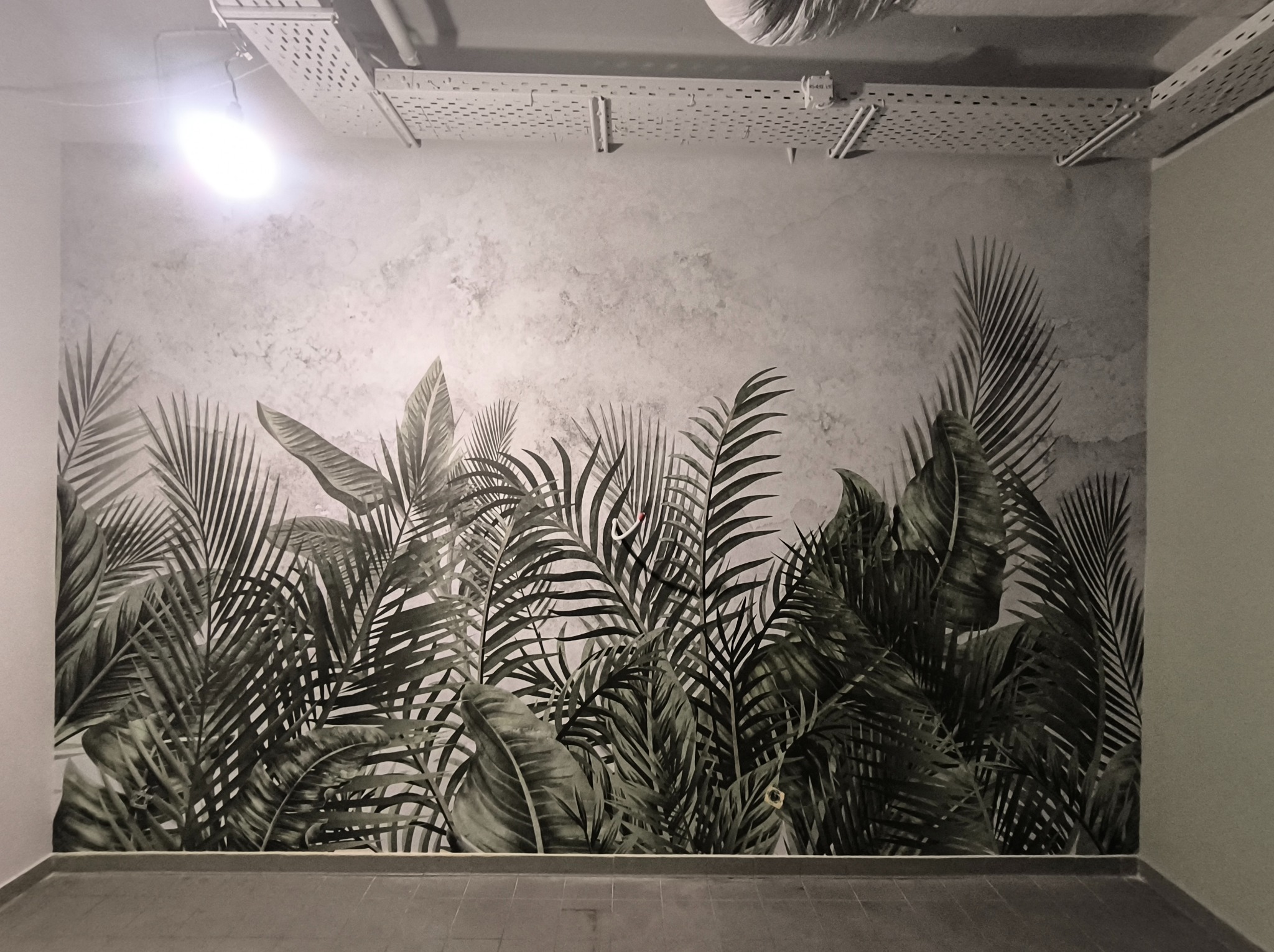 Ściana po dekoracyjnym malowaniu z motywem tropikalnych liści, widoczne instalacje pod sufitem. Szaro-zielona tonacja, nowoczesny design wnętrza.