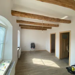 Jasne wnętrze z drewnianymi belkami stropowymi i dwiema parami drzwi z drewnianymi futrynami. Podłoga wyłożona panelami imitującymi drewno.