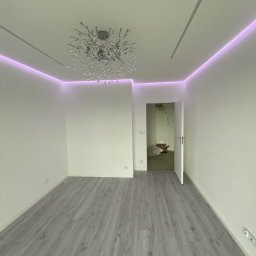 Viktor Yevsiukov - Wykończone wnętrze pokoju w Katowicach z szarą podłogą, białymi ścianami i dekoracyjnym oświetleniem LED oraz żyrandolem. Widok na otwarte drzwi do innego pomieszczenia.