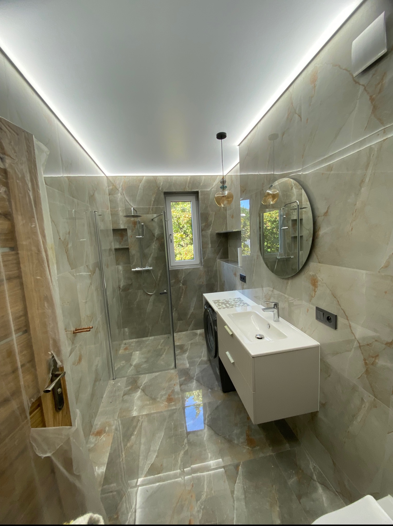 Wykończona łazienka z kabiną prysznicową typu walk-in, umywalką nablatową i marmurowymi płytkami. Widoczne oświetlenie LED podsufitowe i wisząca lampa.