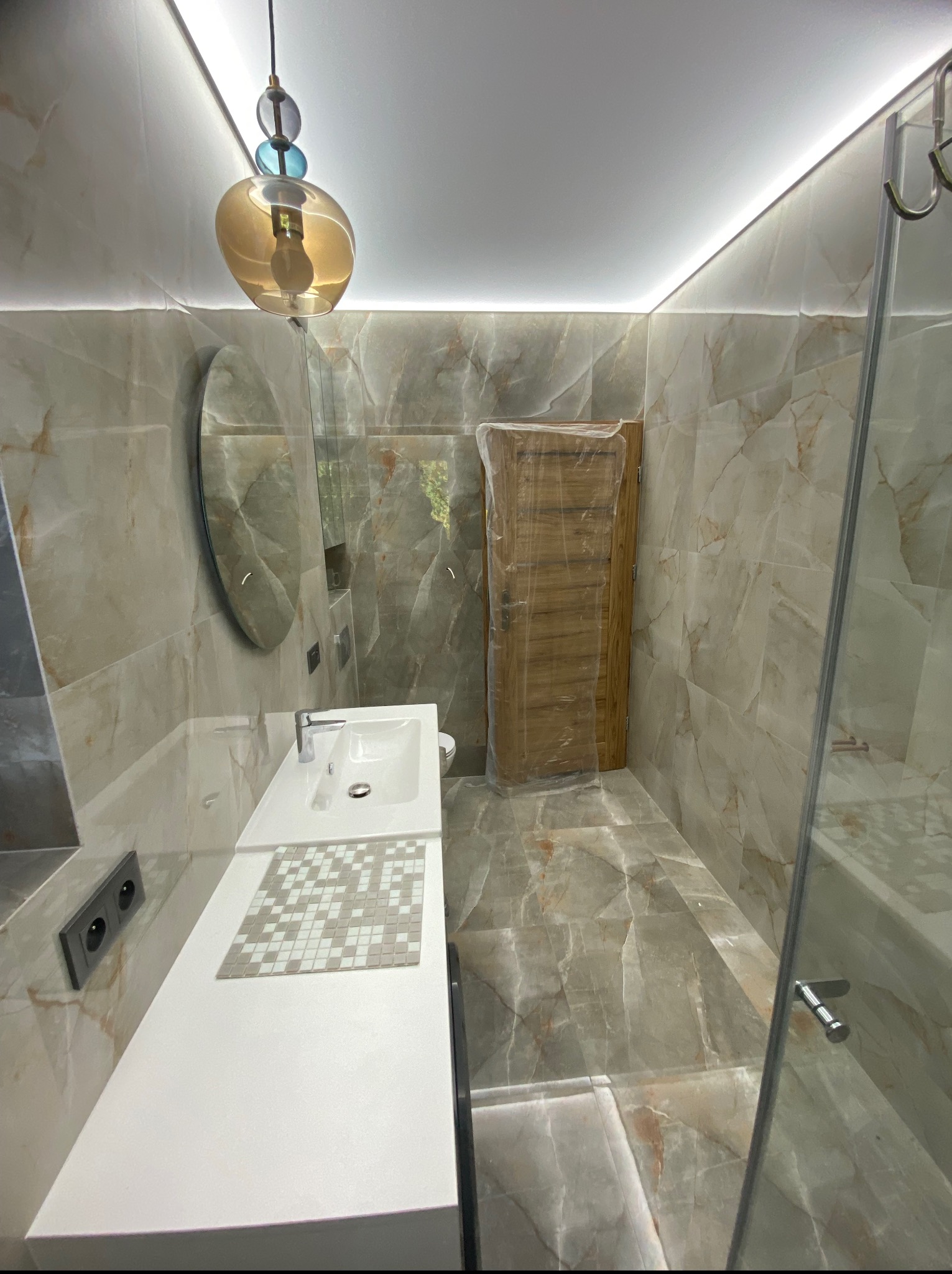 Nowoczesna łazienka po remoncie: marmurowe płytki, biała umywalka z mozaiką, szklana kabina prysznicowa, designerska lampa i podświetlenie LED.