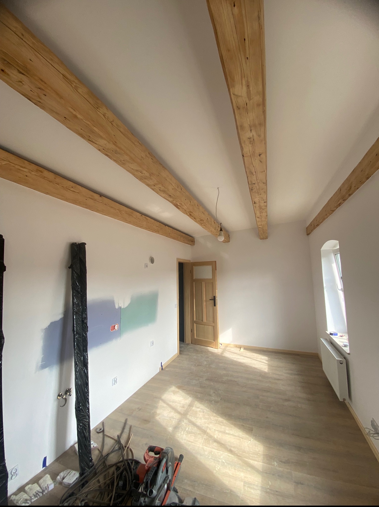 Jasne wnętrze z drewnianymi belkami stropowymi, fragmentami malowania ścian i nową podłogą. Widoczne narzędzia i drzwi z ościeżnicą.