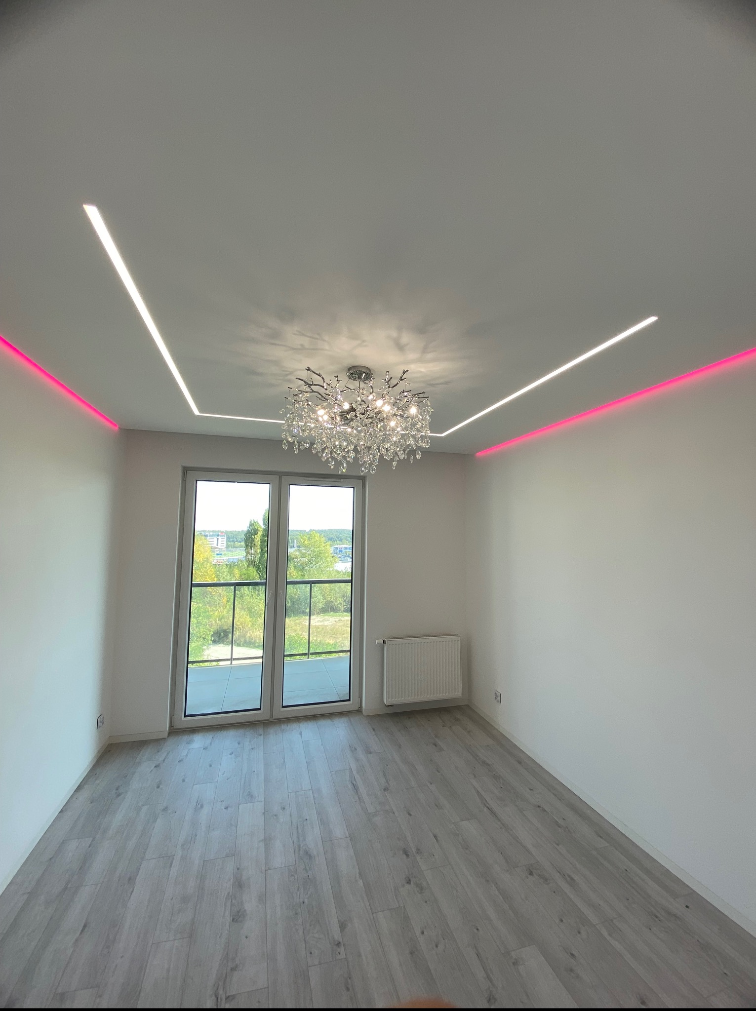 Jasne wnętrze pokoju z panelami podłogowymi, białymi ścianami, grzejnikiem, oknem balkonowym i dekoracyjnym oświetleniem sufitowym LED z żyrandolem.