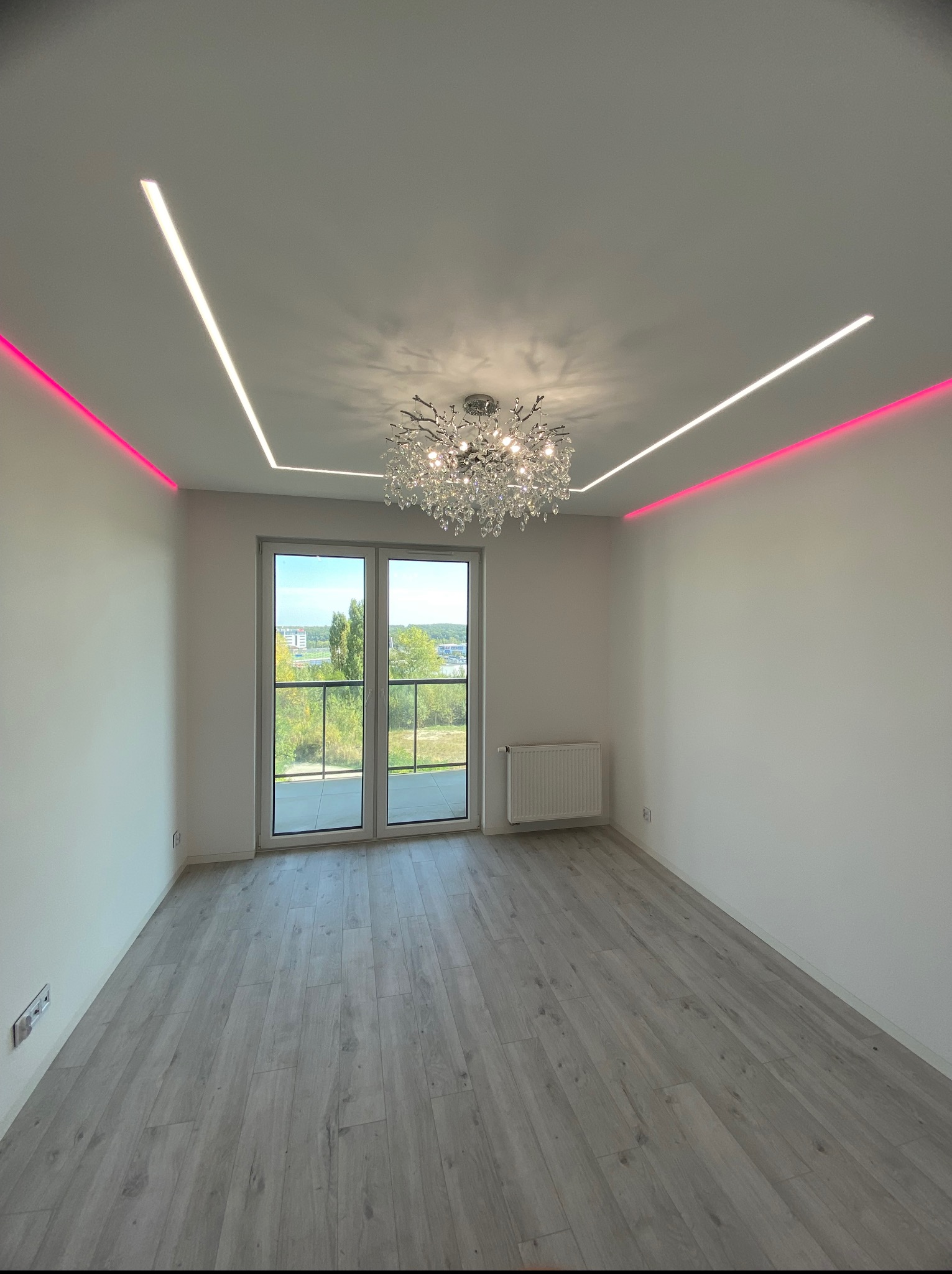 Jasne wnętrze z parkietem, ozdobnym żyrandolem i nowoczesnym oświetleniem LED wzdłuż sufitu. Widok na balkon przez przeszklone drzwi.