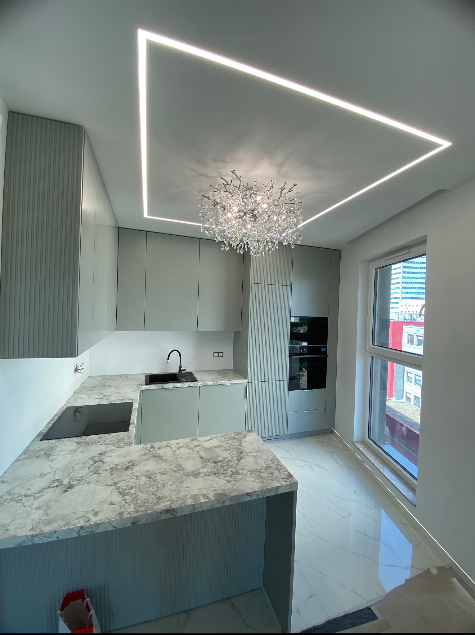 Nowoczesna kuchnia w szarościach z marmurowym blatem, oświetleniem LED na suficie i kryształowym żyrandolem. Widok na miasto przez okno.