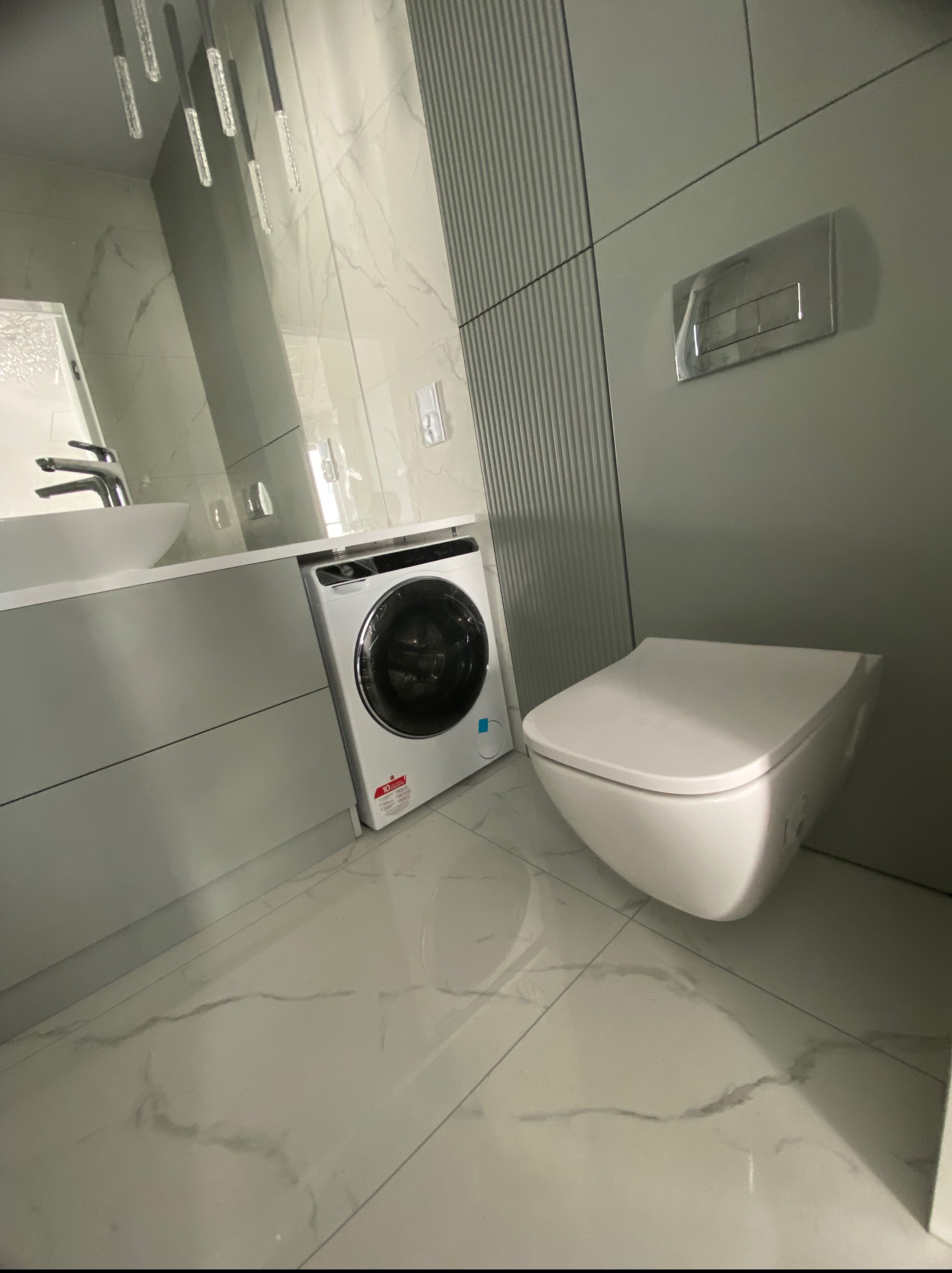 Nowoczesna łazienka w odcieniach szarości z marmurową posadzką, podwieszaną toaletą i pralką pod umywalką. Oświetlenie punktowe i minimalistyczny design.