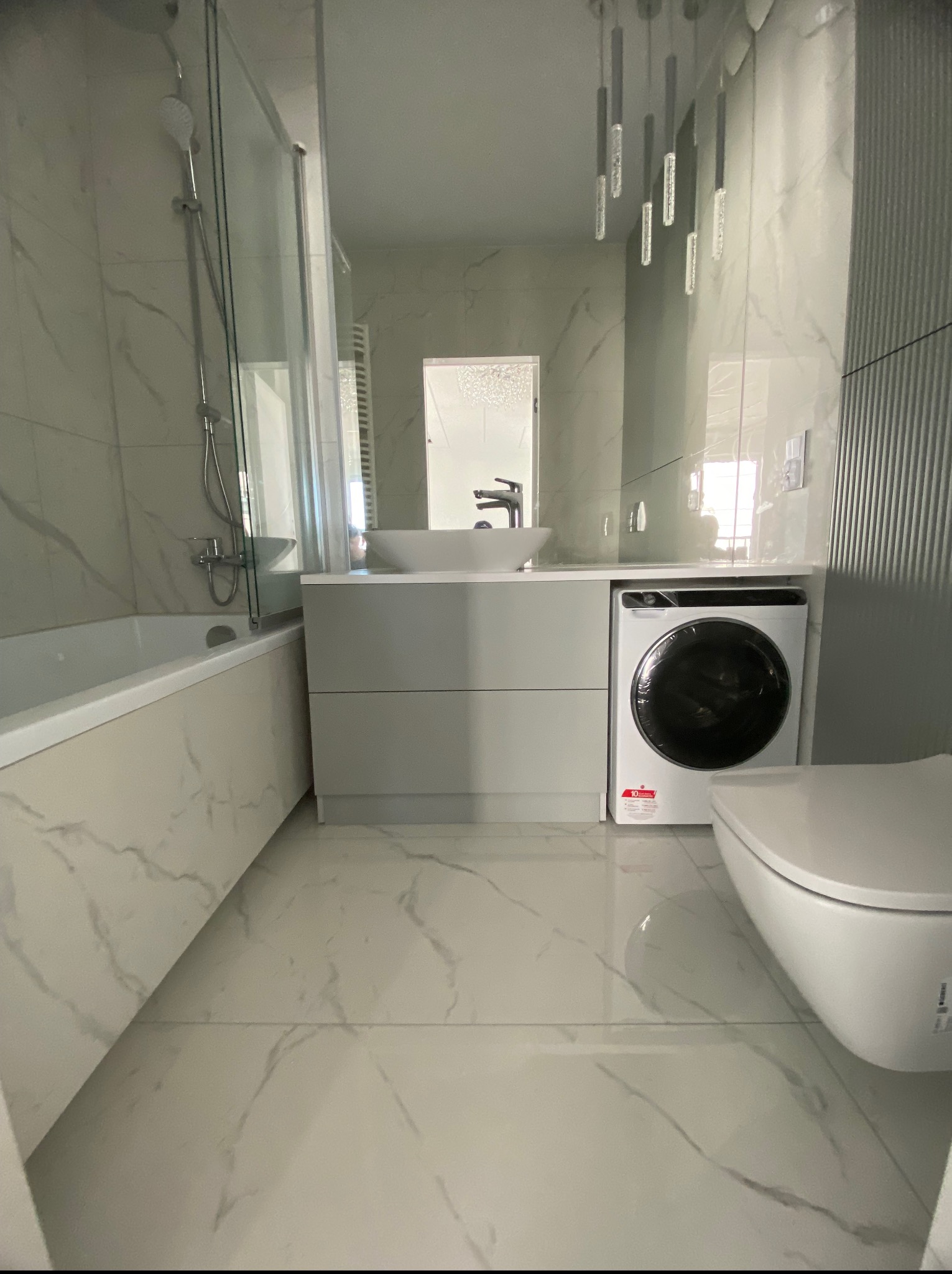 Nowoczesna łazienka z wanną, kabiną prysznicową, umywalką w zabudowie z pralką i toaletą. Białe marmurowe płytki na ścianach i podłodze. Wiszące oświetlenie.