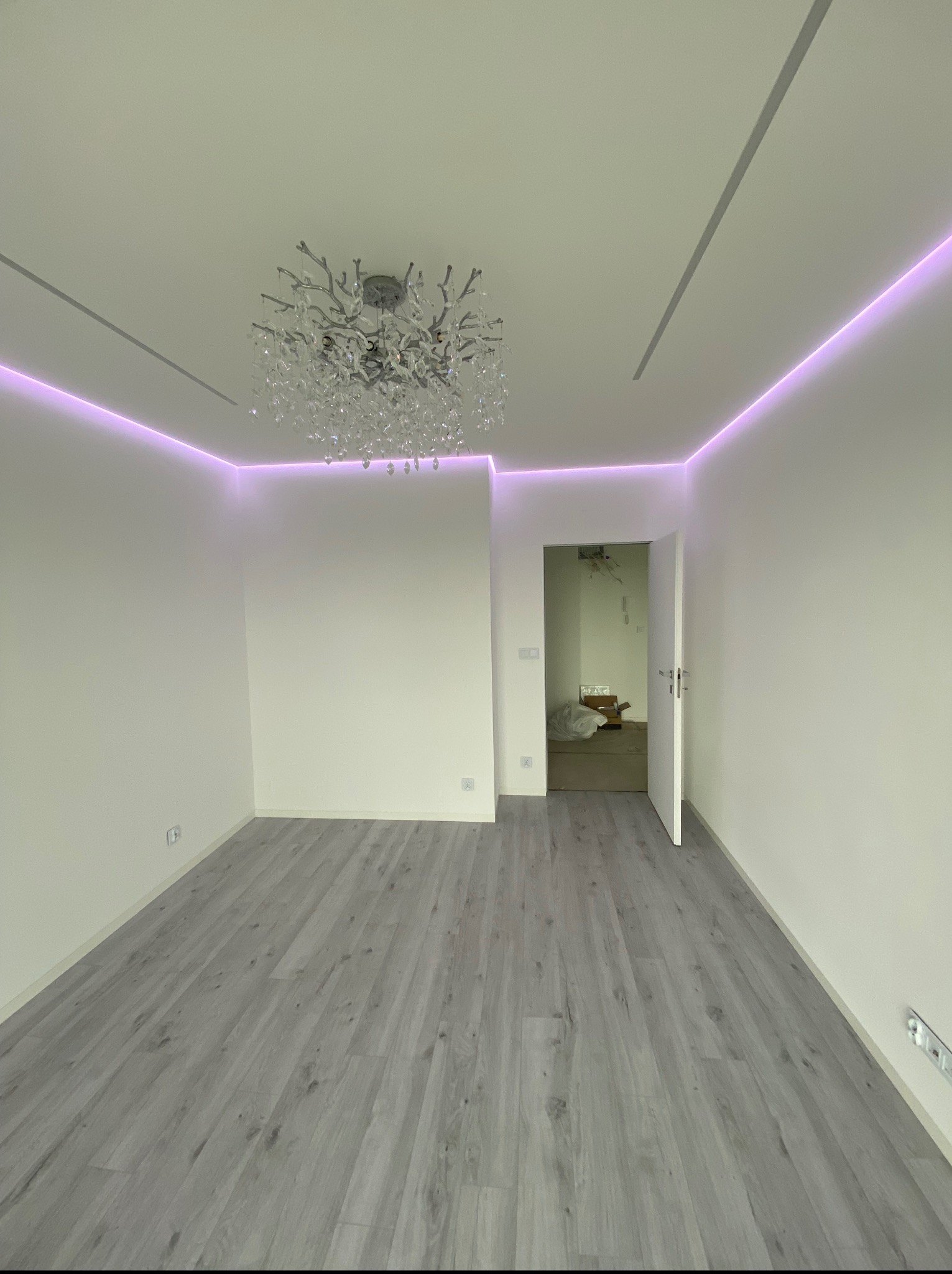 Wykończone wnętrze pokoju w Katowicach z szarą podłogą, białymi ścianami i dekoracyjnym oświetleniem LED oraz żyrandolem. Widok na otwarte drzwi do innego pomieszczenia.