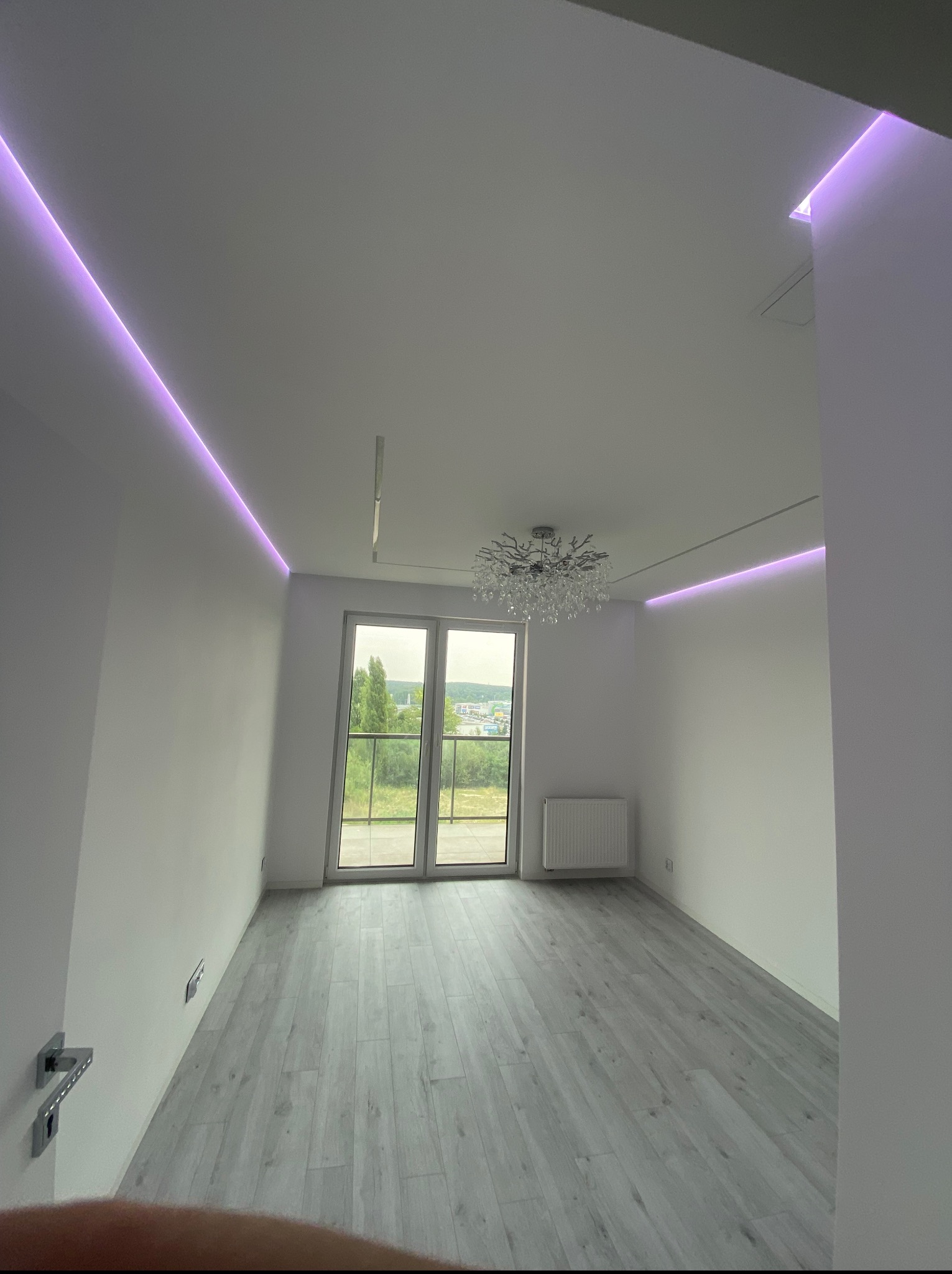 Jasne wnętrze pokoju z szarymi panelami podłogowymi, oświetleniem LED na suficie i żyrandolem. Widok na zewnątrz przez przeszklone drzwi balkonowe.