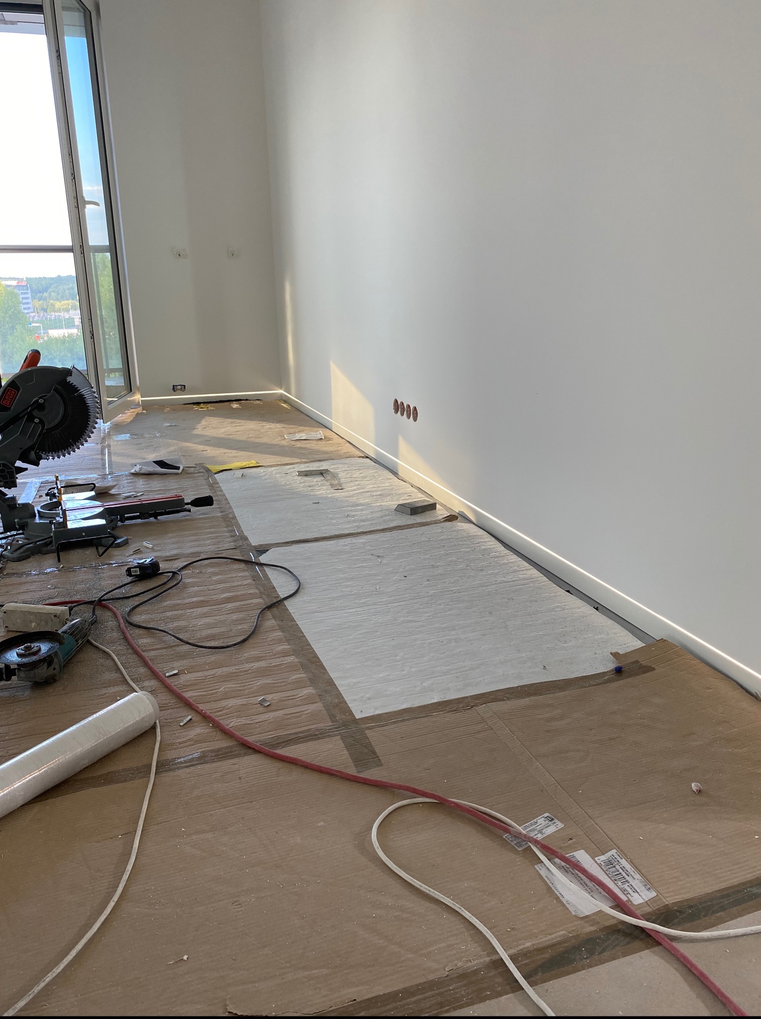 Pokój w trakcie prac wykończeniowych: podłoga zabezpieczona kartonem, widoczne narzędzia (piła, szlifierka), listwy przypodłogowe i instalacja elektryczna.