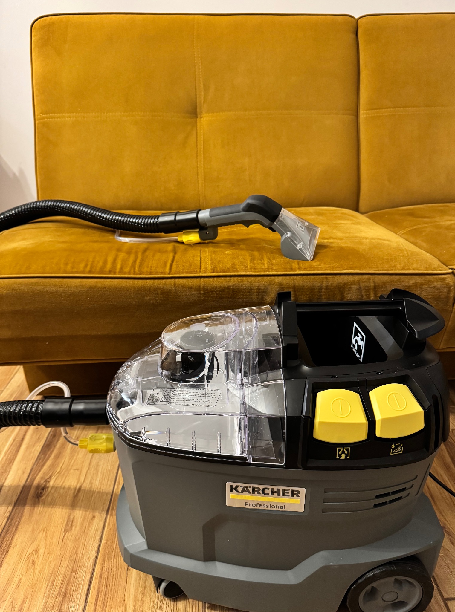 Profesjonalny odkurzacz Kärcher do prania tapicerki, z dyszą na welurowej, musztardowej kanapie. Widoczny wąż ssący i żółte przyciski.