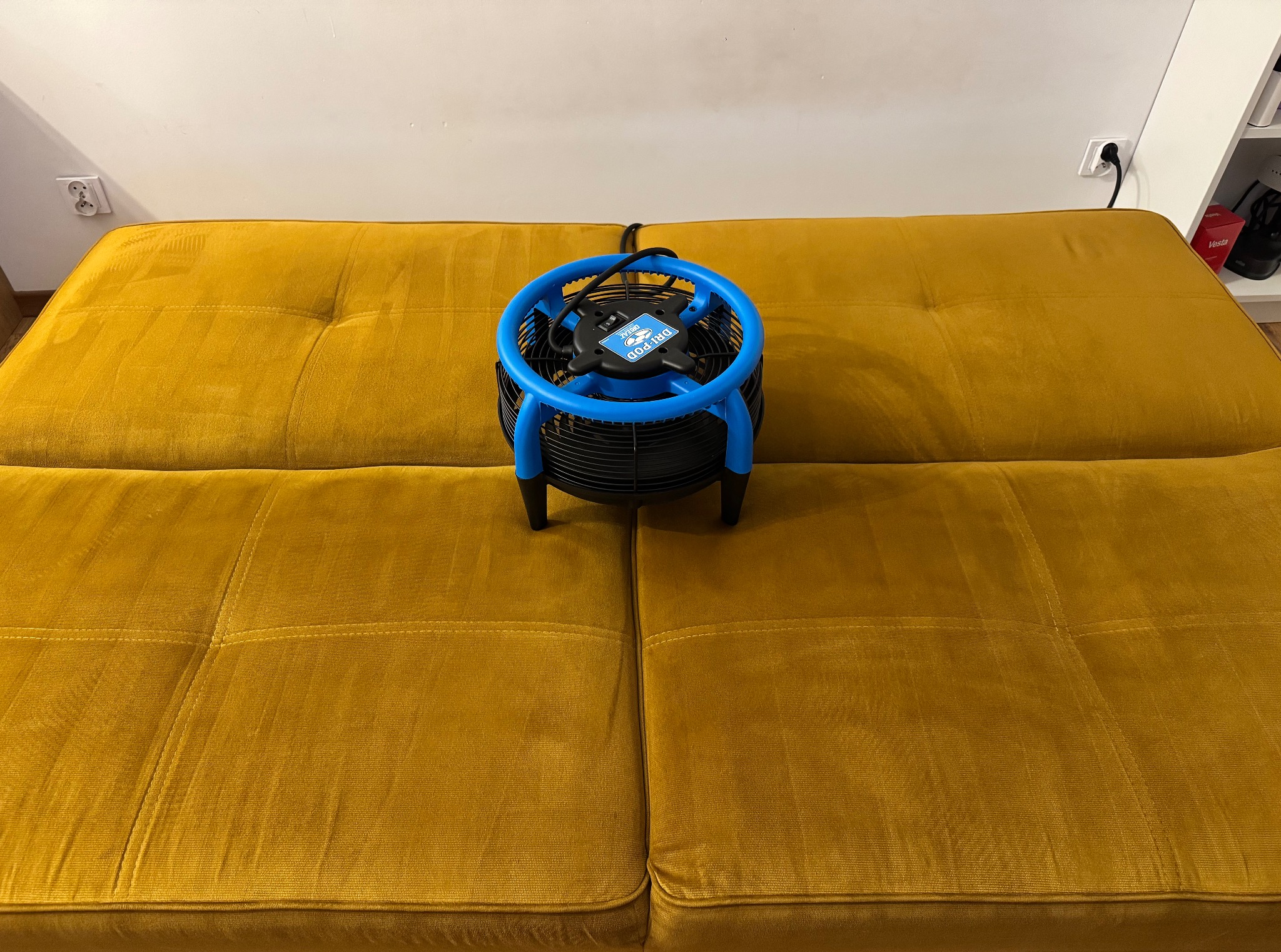 Żółta kanapa z wentylatorem Dri-Pod do suszenia tapicerki. Urządzenie stoi na środku kanapy, widoczne gniazdko elektryczne w tle.