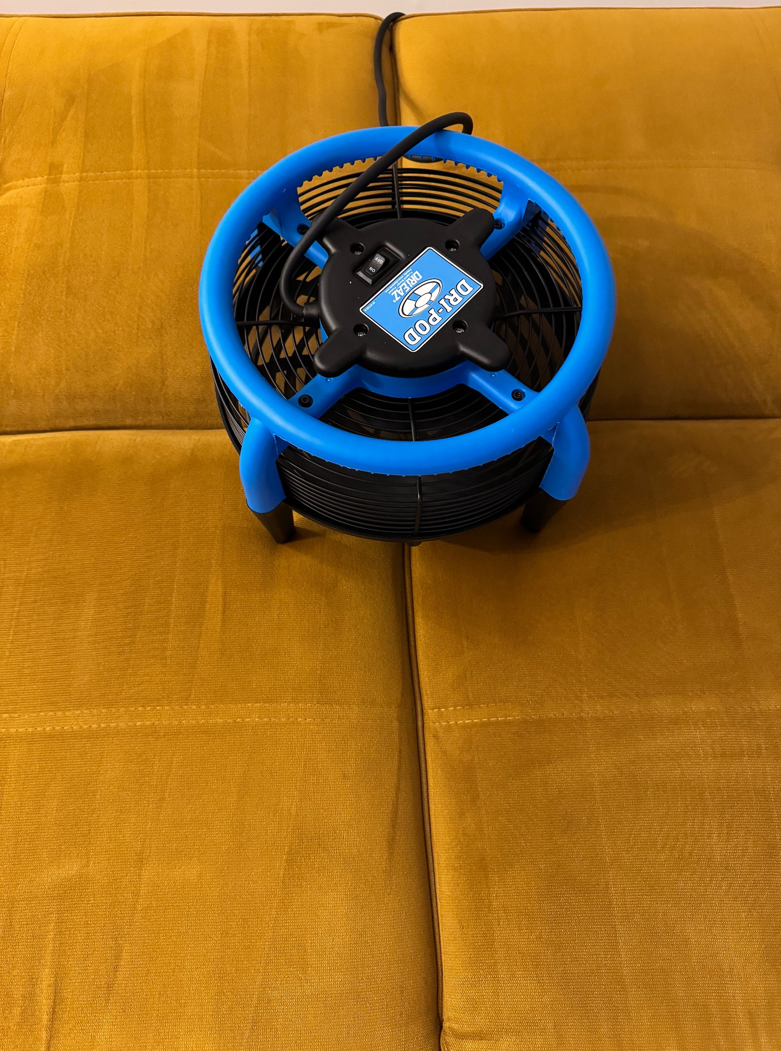 Niebiesko-czarny wentylator DRY-POD na musztardowej kanapie z weluru. Sprzęt do osuszania po praniu tapicerki. Widok z góry, oświetlenie naturalne.