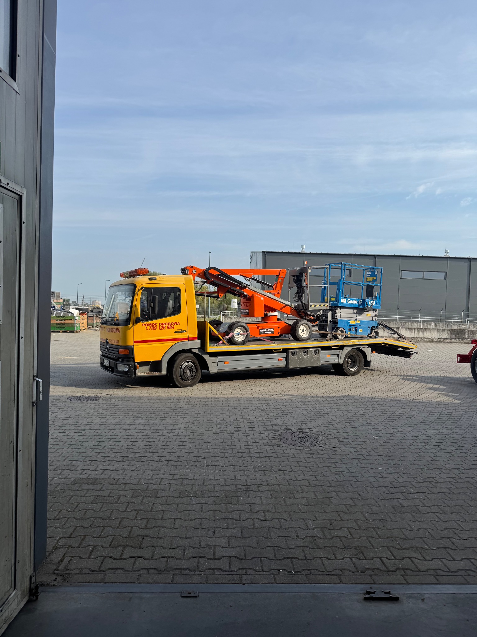 Żółta laweta przewozi podnośniki koszowe na tle szarego budynku. Widoczne pomarańczowe i niebieskie podnośniki.