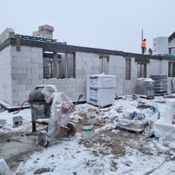 P-SOFT DANIEL PIOTROWSKI - Budowa domu w zimowej scenerii: ściany z betonu komórkowego, widoczna betoniarka, palety z materiałami budowlanymi i pracownicy na dachu. Etap murowania ścian.