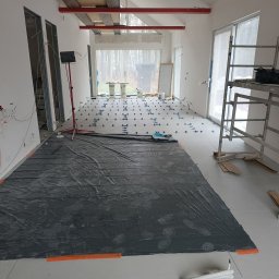 P-SOFT DANIEL PIOTROWSKI - Układanie płytek podłogowych w trakcie remontu w Warszawie. Widoczne poziomowanie i folia ochronna. W tle okno z widokiem na drzewa i rusztowanie.