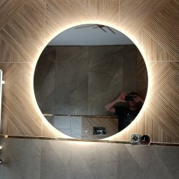 P-SOFT DANIEL PIOTROWSKI - Wykończona łazienka z okrągłym lustrem LED i grzejnikiem drabinkowym. Na ścianach płytki imitujące drewno i kamień. Widoczne gniazdka elektryczne i odbicie fotografa.