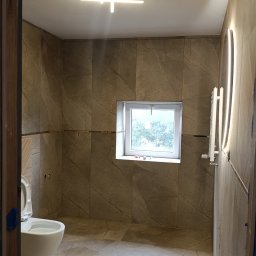 P-SOFT DANIEL PIOTROWSKI - Wykończona łazienka z beżowymi płytkami imitującymi marmur, nowoczesnym oświetleniem sufitowym i grzejnikiem drabinkowym. Widok zza framugi drzwi.