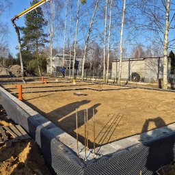 P-SOFT DANIEL PIOTROWSKI - Fundamenty domu jednorodzinnego w trakcie budowy, widoczne zbrojenia, izolacja pionowa i rury kanalizacyjne. W tle drzewa i budynki.