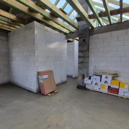 Wnętrze surowego domu w trakcie budowy: ściany z białych bloczków, drewniana konstrukcja dachu, karton Velux i paleta z materiałami budowlanymi. Widok z poziomu podłogi.