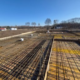 Zbrojenie płyty fundamentowej domu jednorodzinnego, widok z góry na siatkę z prętów stalowych i szalunki. W tle budynki i drzewa pod błękitnym niebem.