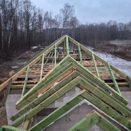 P-SOFT DANIEL PIOTROWSKI - Konstrukcja dachu z impregnowanego drewna na ścianach z betonu komórkowego. Widok z góry na budowę domu w trakcie wznoszenia. W tle las i pochmurne niebo.