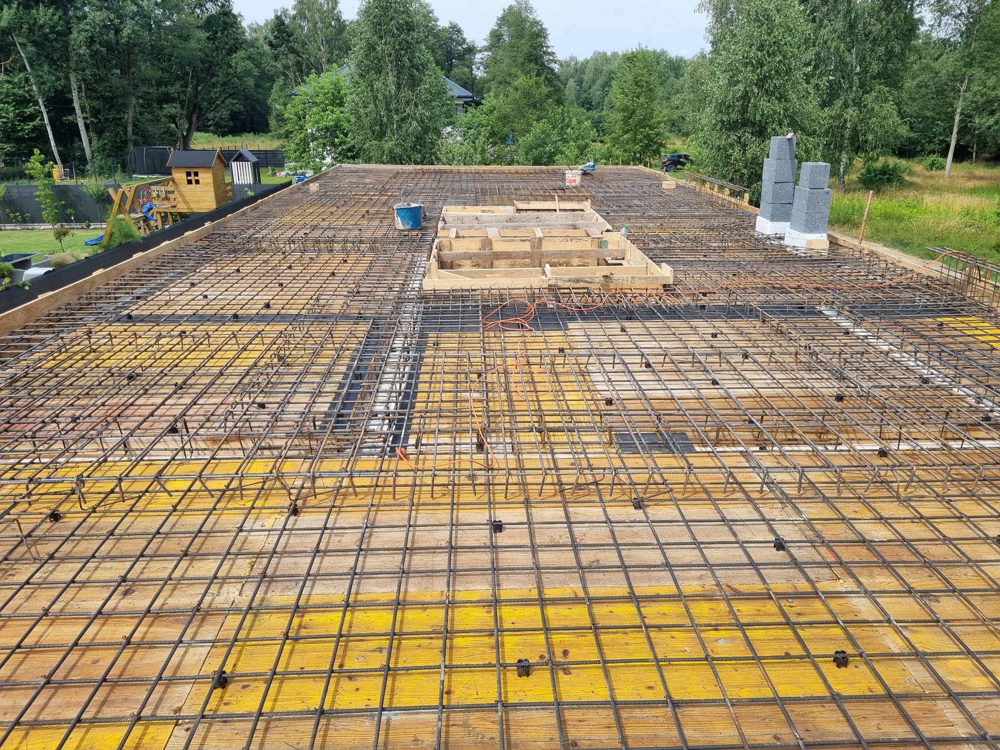 Zbrojenie płyty fundamentowej na żółtym szalunku z widocznym wykopem pod schody i kominem. W tle zieleń drzew i zabudowa. Prace budowlane w toku.