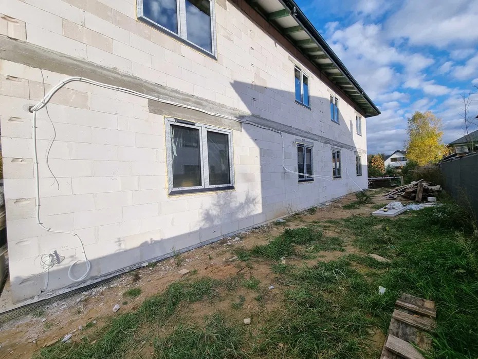 Ściana domu z białego betonu komórkowego w trakcie budowy, widoczne okna z folią ochronną, instalacja elektryczna na zewnątrz, trawa i niebo w tle.