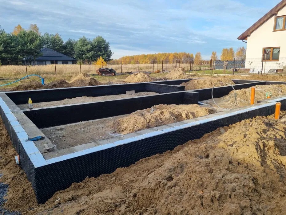 Zabezpieczone fundamenty domu jednorodzinnego z izolacją pionową i poziomą, widoczne rury kanalizacyjne, teren przygotowany pod dalsze prace budowlane.