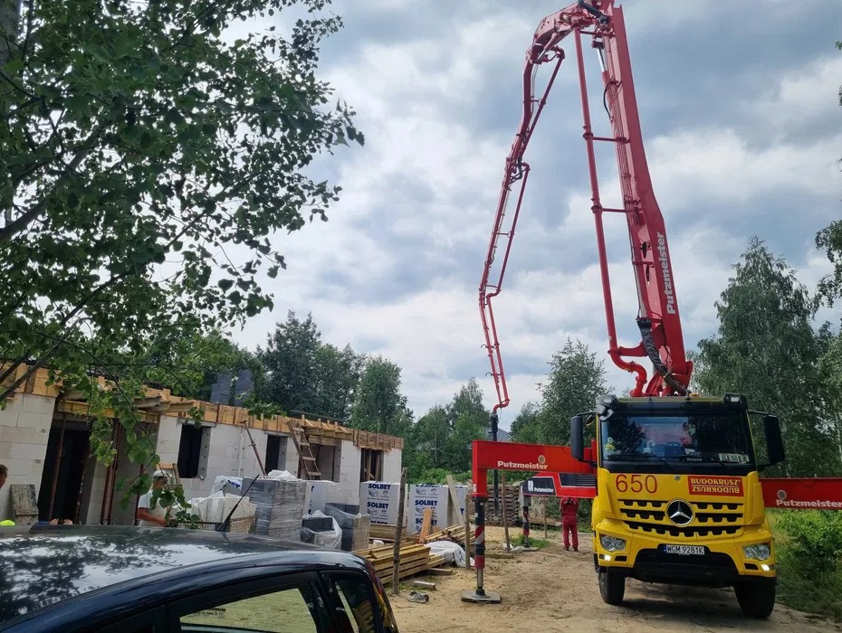 Żółta betoniarka Putzmeister z wysięgnikiem na tle wznoszonego domu z białego bloczka Solbet, częściowo zadaszonego drewnianą konstrukcją. W tle drzewa i pochmurne niebo.