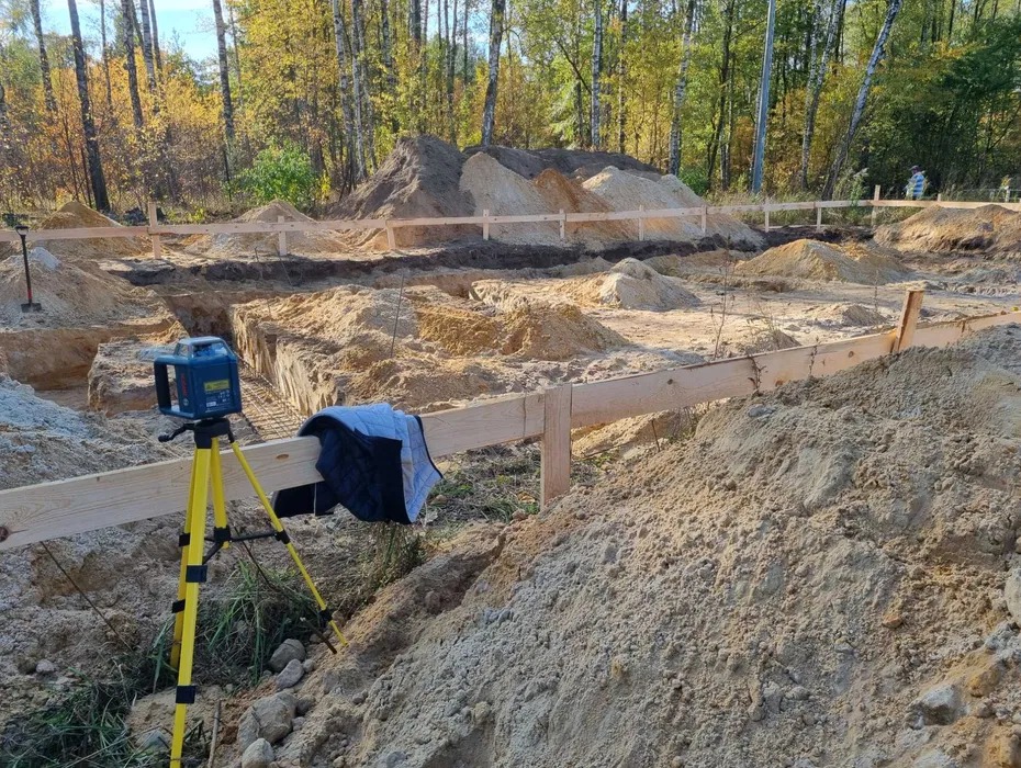 Wykonane wykopy pod fundamenty z widocznym zbrojeniem, drewnianym szalunkiem i niwelatorem laserowym na statywie. W tle drzewa.
