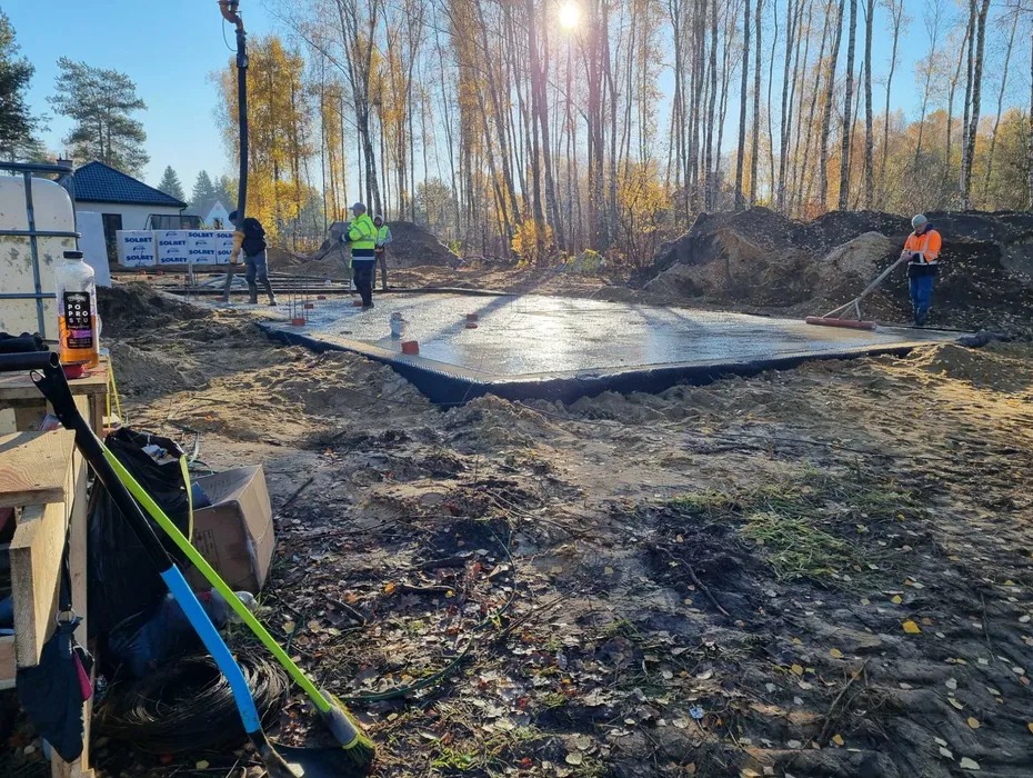 Wylewka fundamentowa w trakcie realizacji. Pracownicy w odblaskowych ubraniach rozprowadzają beton. W tle drzewa i budynek. Widoczne narzędzia i materiały budowlane.