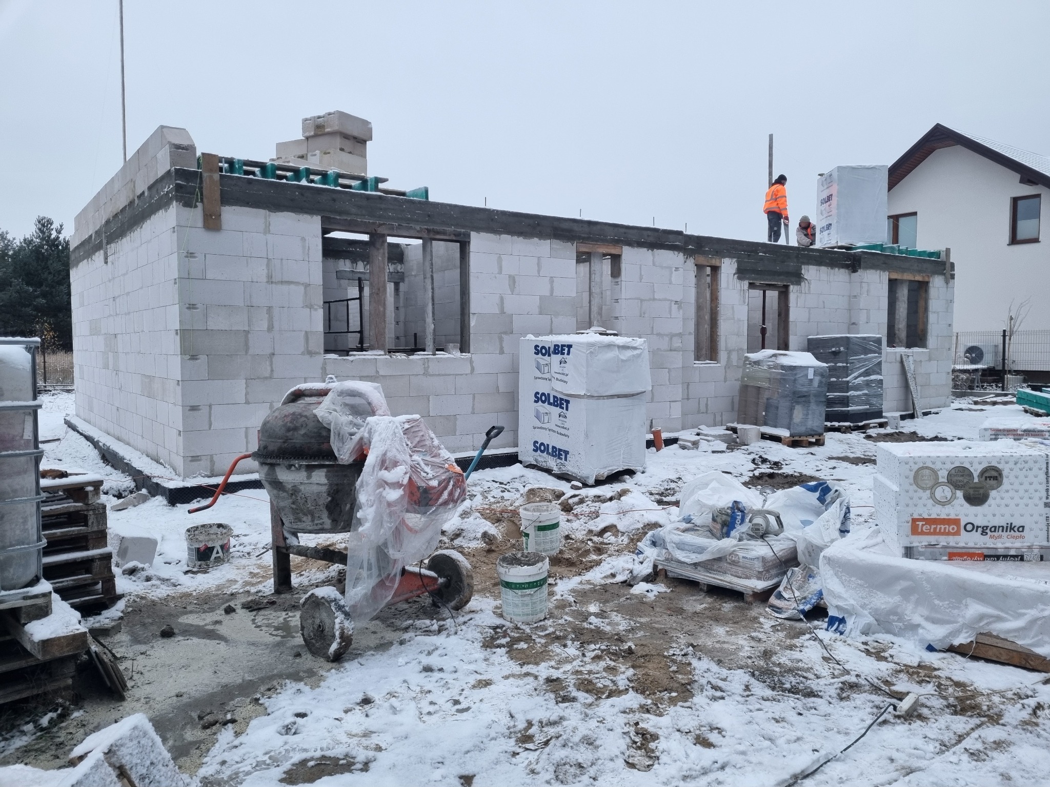 Budowa domu w zimowej scenerii: ściany z betonu komórkowego, widoczna betoniarka, palety z materiałami budowlanymi i pracownicy na dachu. Etap murowania ścian.
