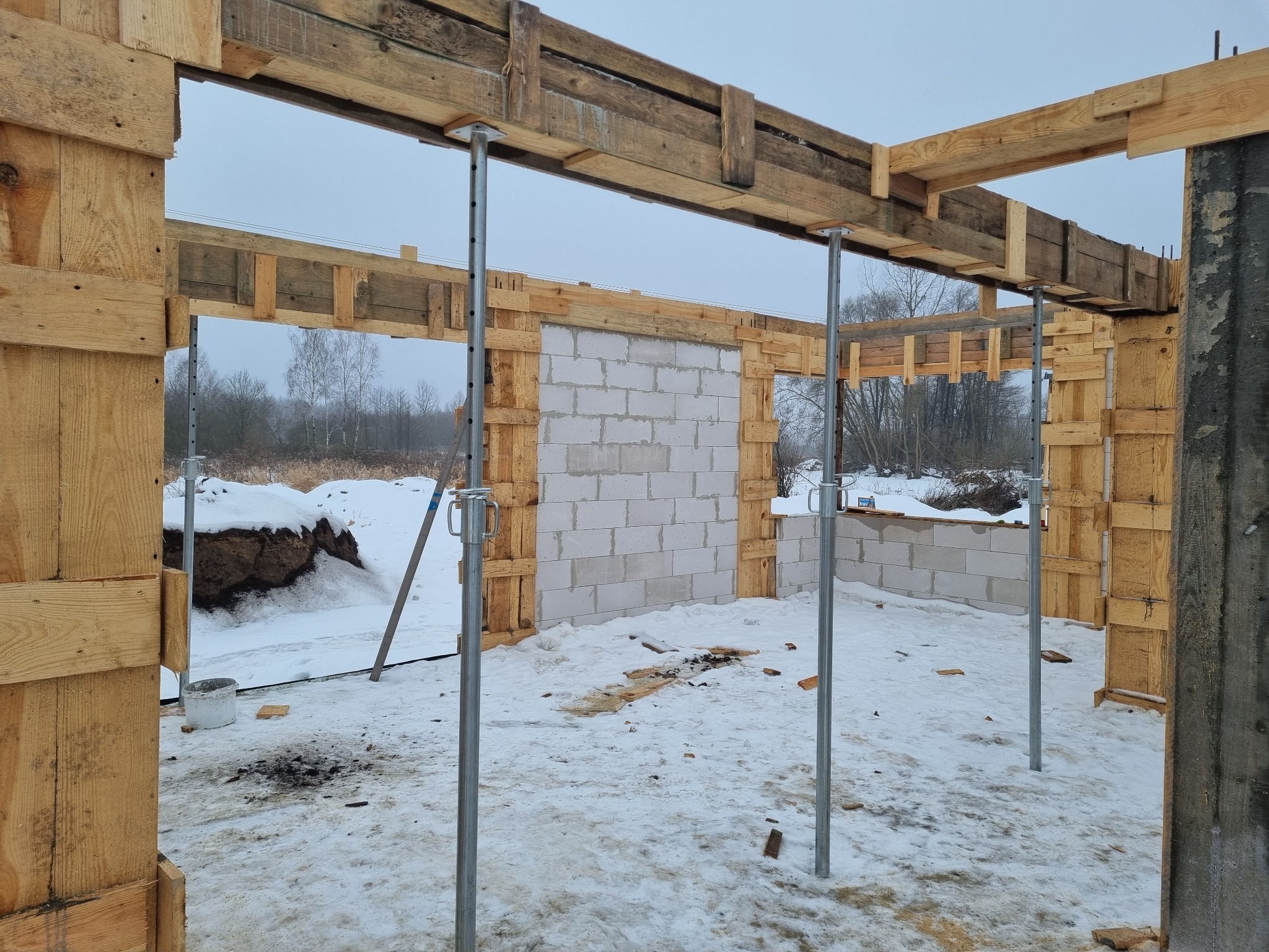 Wnętrze surowego stanu domu w zimowej scenerii. Widoczne drewniane szalunki, ściany z bloczków, metalowe podpory i śnieg na zewnątrz. Etap budowy z widocznym krajobrazem.