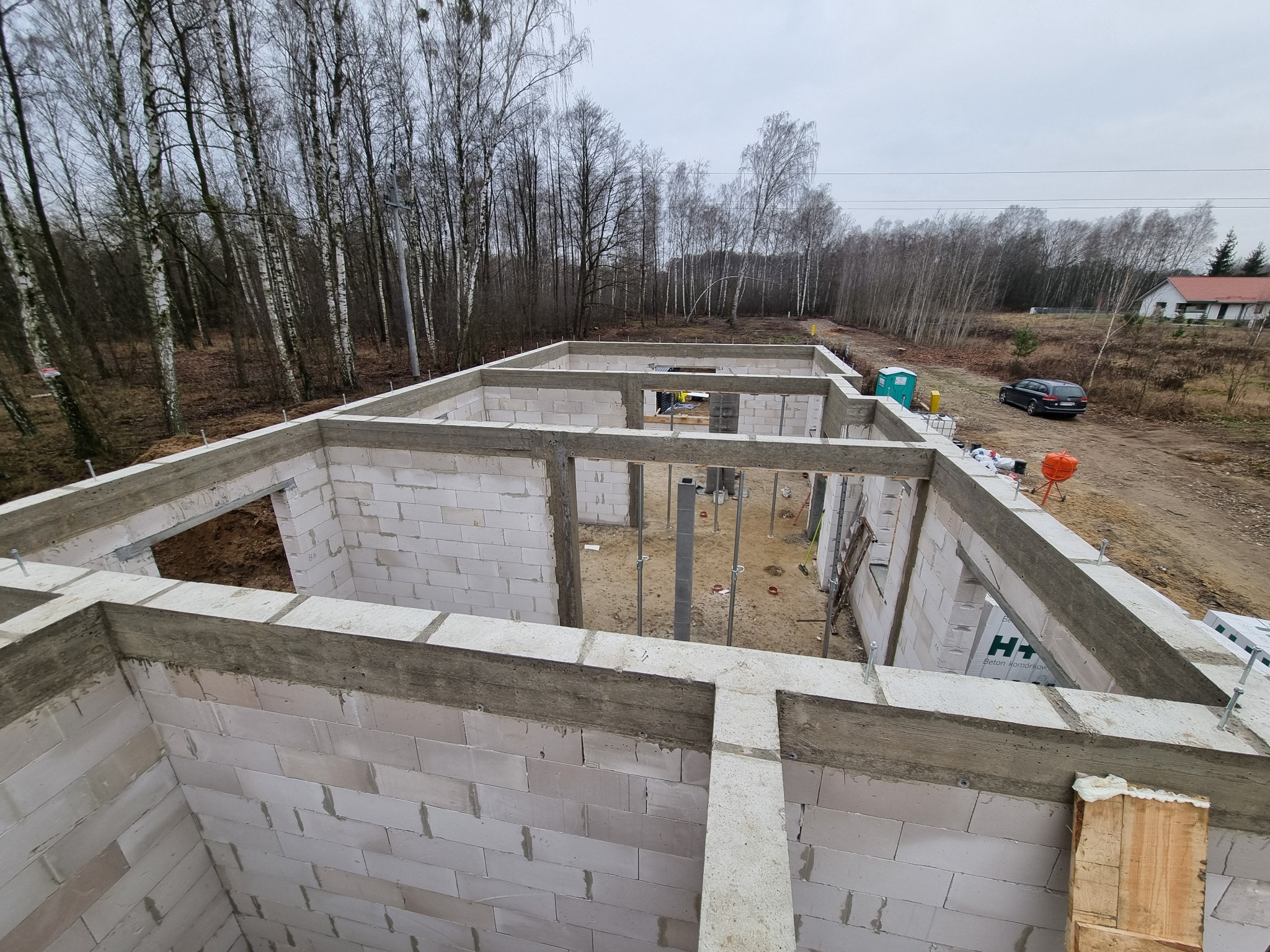 Surowy stan domu z betonu komórkowego, widok z góry na ściany nośne i fundamenty, na zewnątrz drzewa i częściowo widoczny dom z czerwonym dachem.