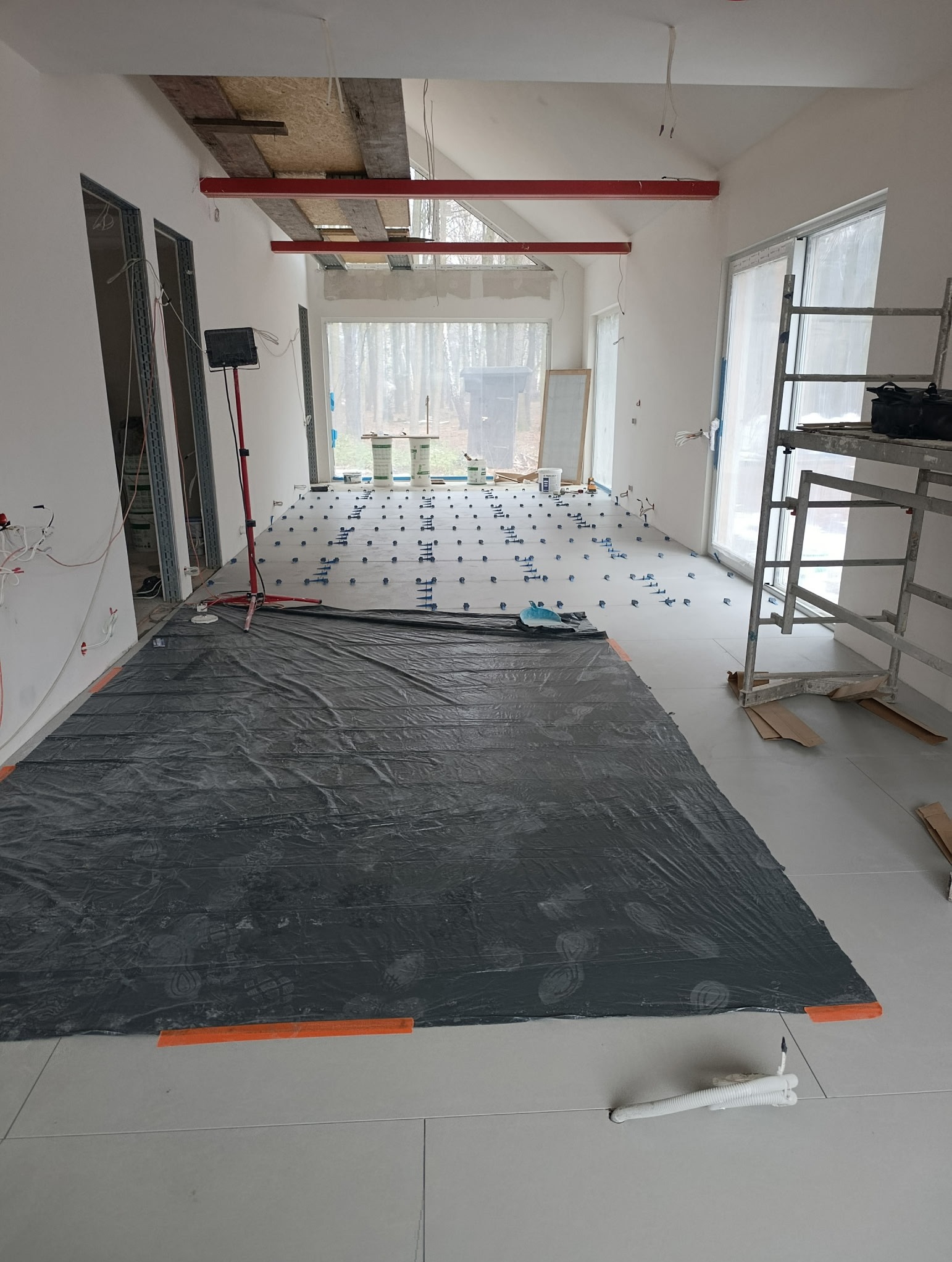 Układanie płytek podłogowych w trakcie remontu w Warszawie. Widoczne poziomowanie i folia ochronna. W tle okno z widokiem na drzewa i rusztowanie.