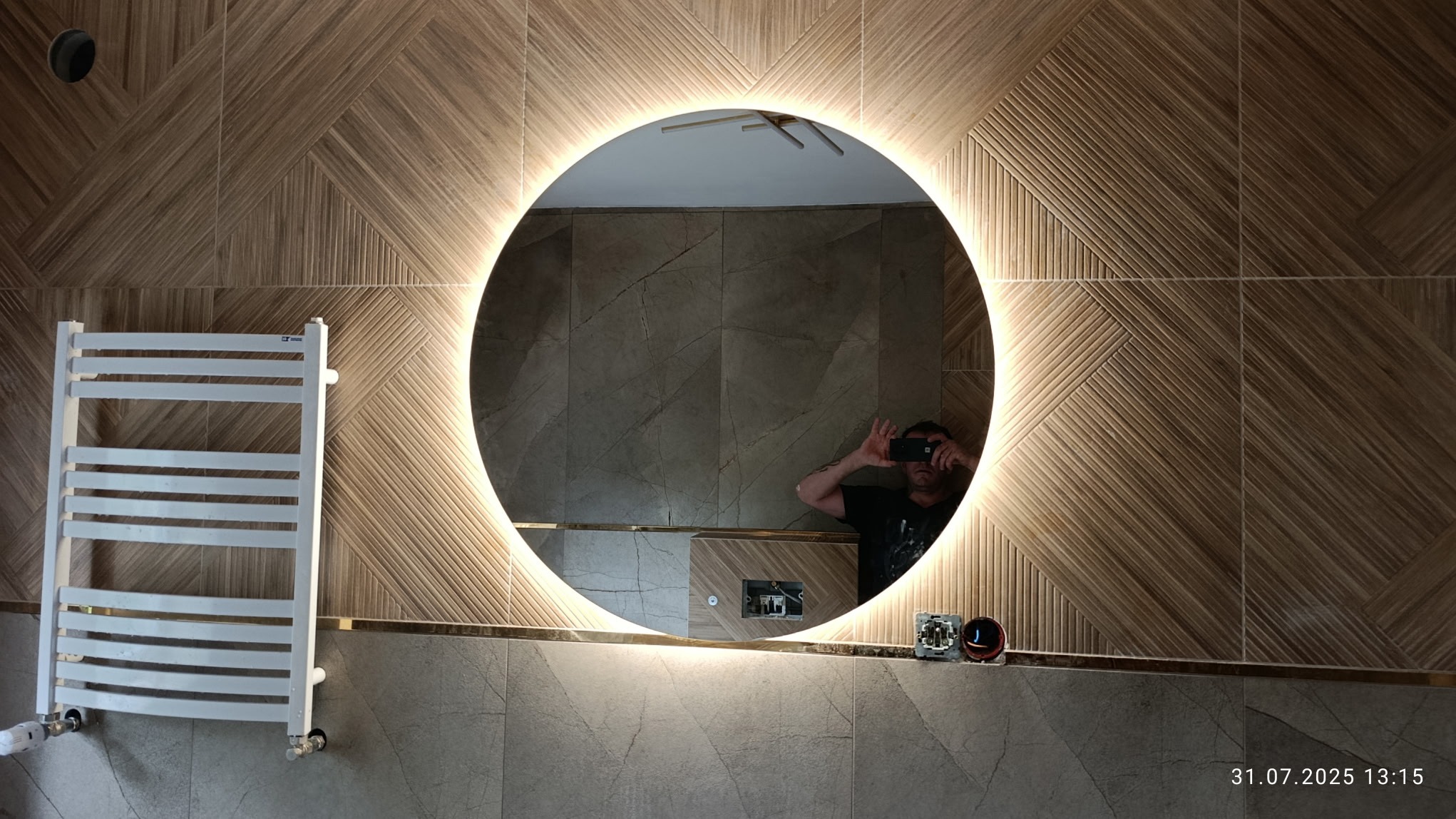 Wykończona łazienka z okrągłym lustrem LED i grzejnikiem drabinkowym. Na ścianach płytki imitujące drewno i kamień. Widoczne gniazdka elektryczne i odbicie fotografa.