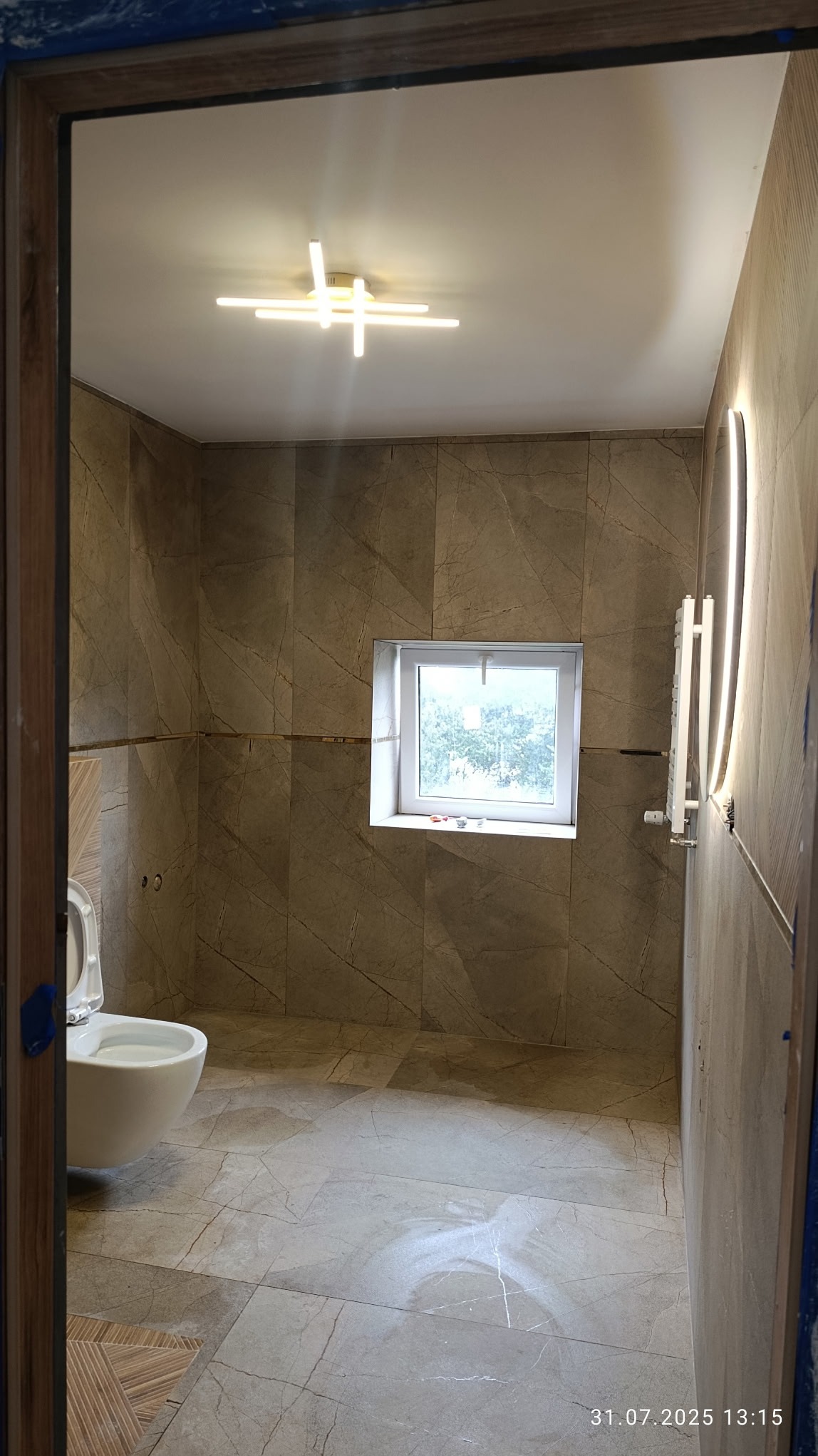 Wykończona łazienka z beżowymi płytkami imitującymi marmur, nowoczesnym oświetleniem sufitowym i grzejnikiem drabinkowym. Widok zza framugi drzwi.