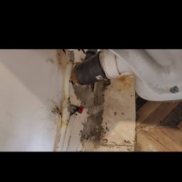 Złota rączka 24h - Niefachowo wykonane podłączenie odpływu toalety. Widoczne zacieki, rdza i uszkodzenia ściany. Elementy hydrauliczne w nieestetycznym stanie.