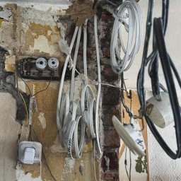 Złota rączka 24h - Niefunkcjonująca instalacja elektryczna z przewodami, puszką bezpiecznikową i gniazdkiem na tle zniszczonej ściany w trakcie remontu. Widoczne kable i fragment ceglanej ściany.