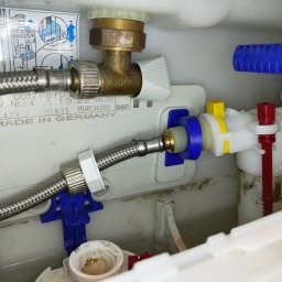 Złota rączka 24h - Wnętrze spłuczki WC z widocznymi elementami instalacji wodnej: zawór Schell, wężyk w oplocie stalowym, elementy z tworzywa w kolorach białym, niebieskim i żółtym. Widoczne instrukcje.