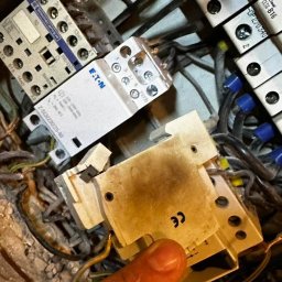 Złota rączka 24h - Uszkodzony stycznik EATON z widocznymi śladami przepalenia, w tle inne elementy instalacji elektrycznej i przewody. Palec wskazujący na uszkodzenie.