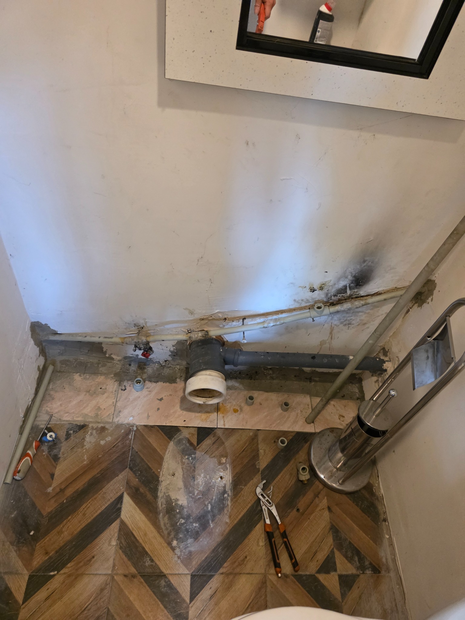 Remont łazienki: widoczne rury kanalizacyjne, narzędzia (klucz nastawny) na podłodze z wzorem jodełki. Fragment ściany z śladami po instalacji. Widok z góry.