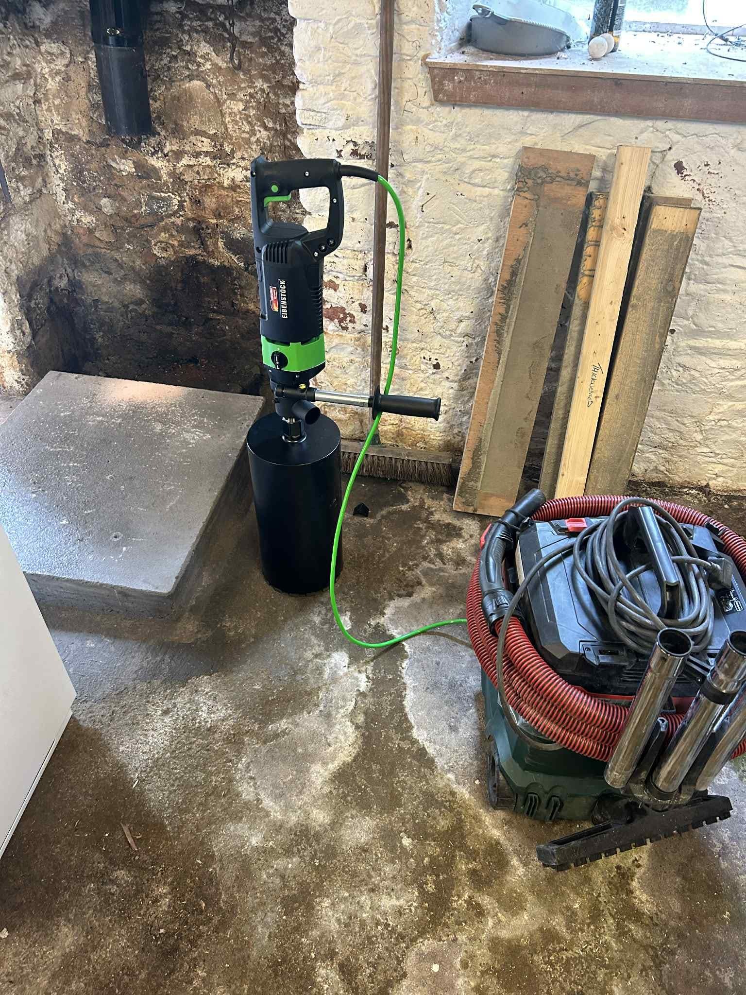 Wiertnica Eibenstock z podłączonym odkurzaczem przemysłowym na wilgotnej posadzce w piwnicy, obok betonowego stopnia i drewnianych desek opartych o ścianę.