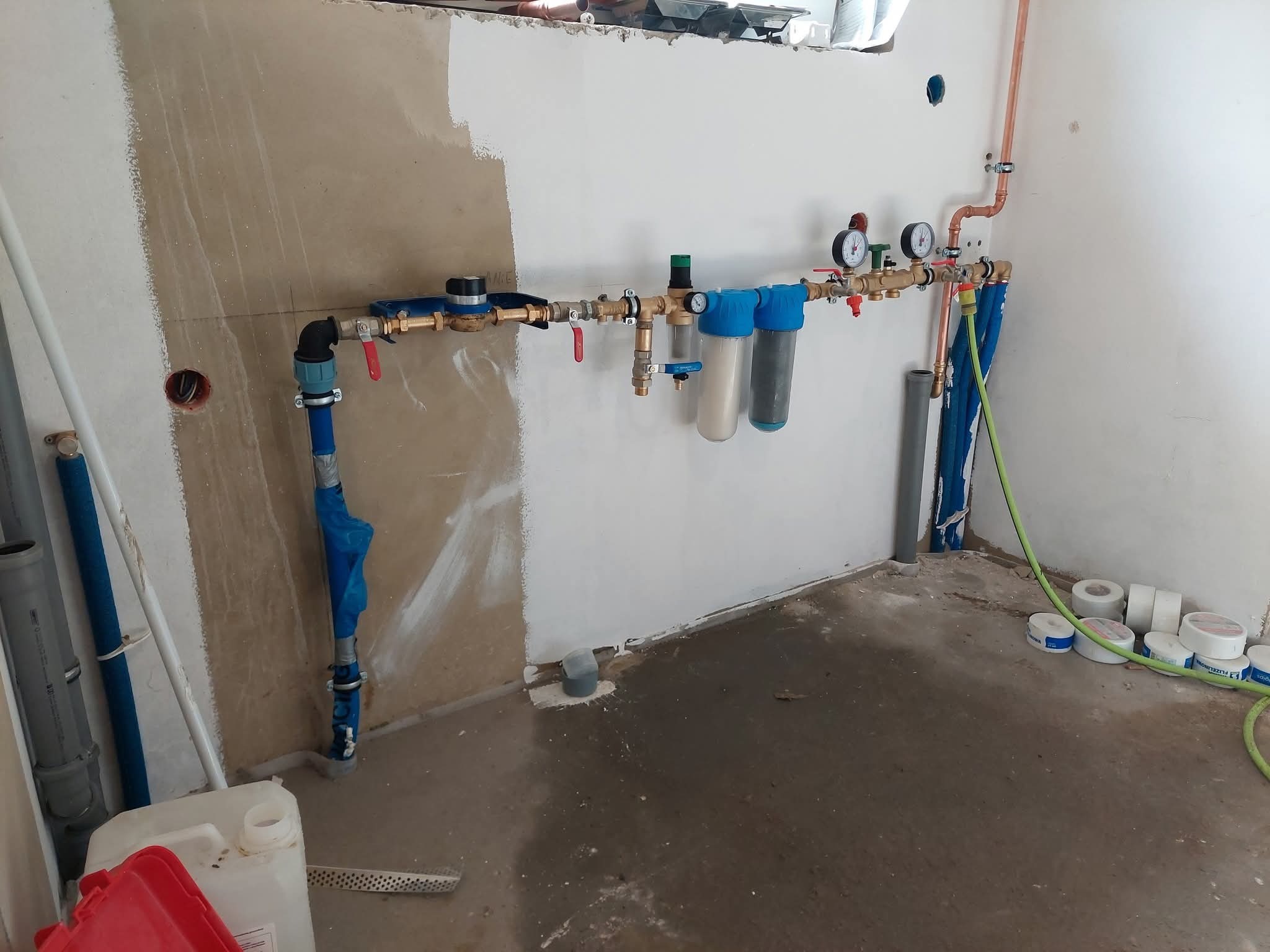 Instalacja wodna z filtrami, manometrami i zaworami, zamontowana na ścianie w surowym stanie. Widoczne rury miedziane i plastikowe, taśmy uszczelniające na podłodze.