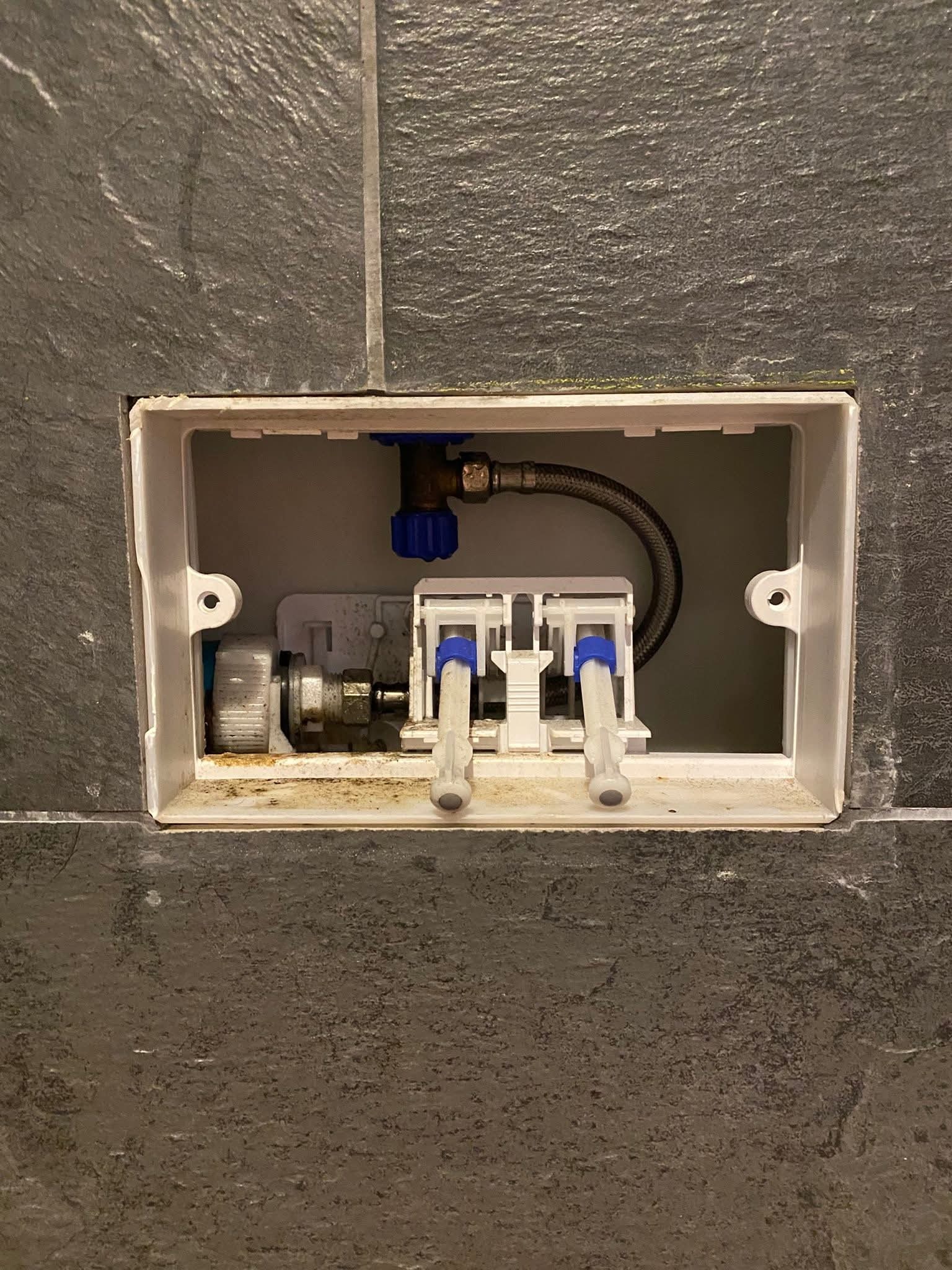 Podłączenie do spłuczki podtynkowej w obudowie z szarej płytki imitującej kamień. Widoczny zawór, wężyk i elementy mocujące. Ujęcie z bliska, na wprost.