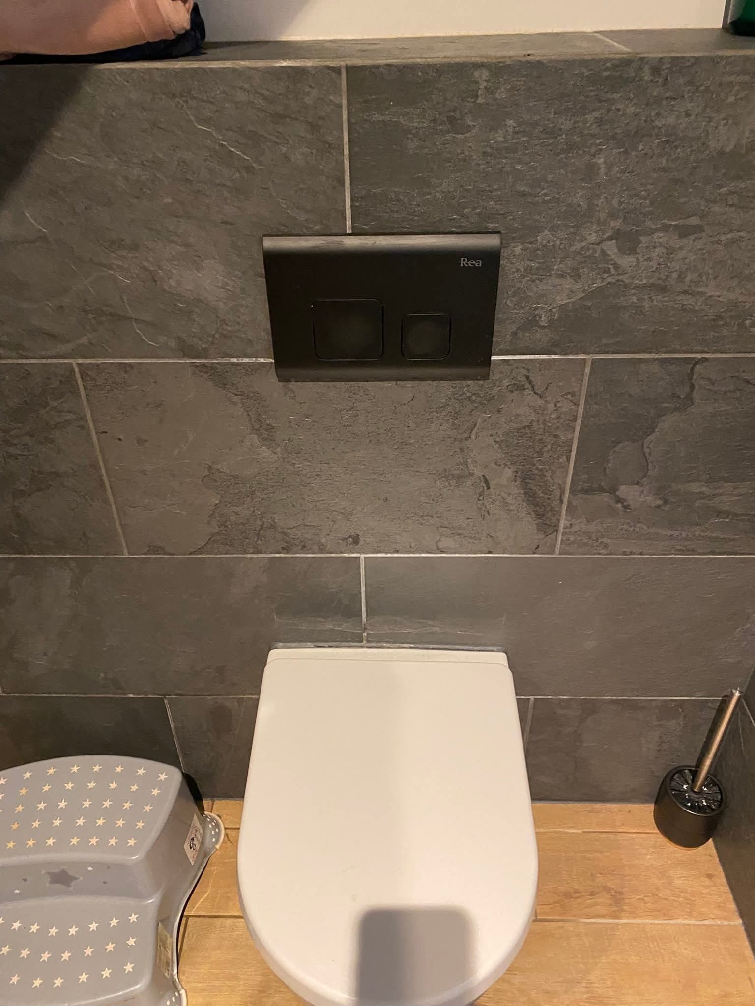 Nowoczesna toaleta wisząca z szarą, kamienną okładziną ścienną, czarnym panelem spłukującym Rea i drewnianą podłogą. Widoczny stołek dla dziecka.